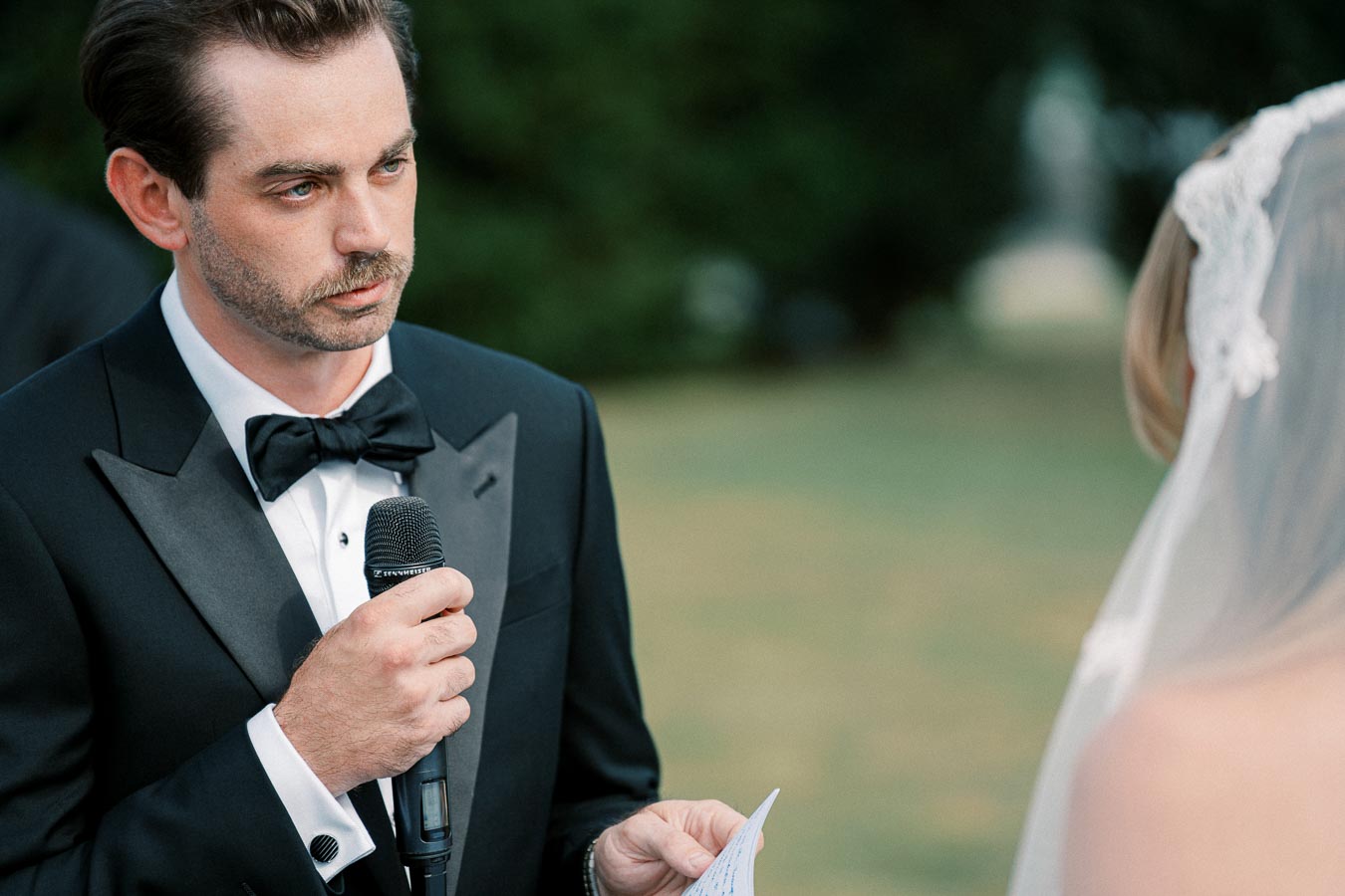 Groom in a black tuxedo holding a microphone, speaking during an outdoor wedding ceremony, with a bride in a white veil in the foreground.