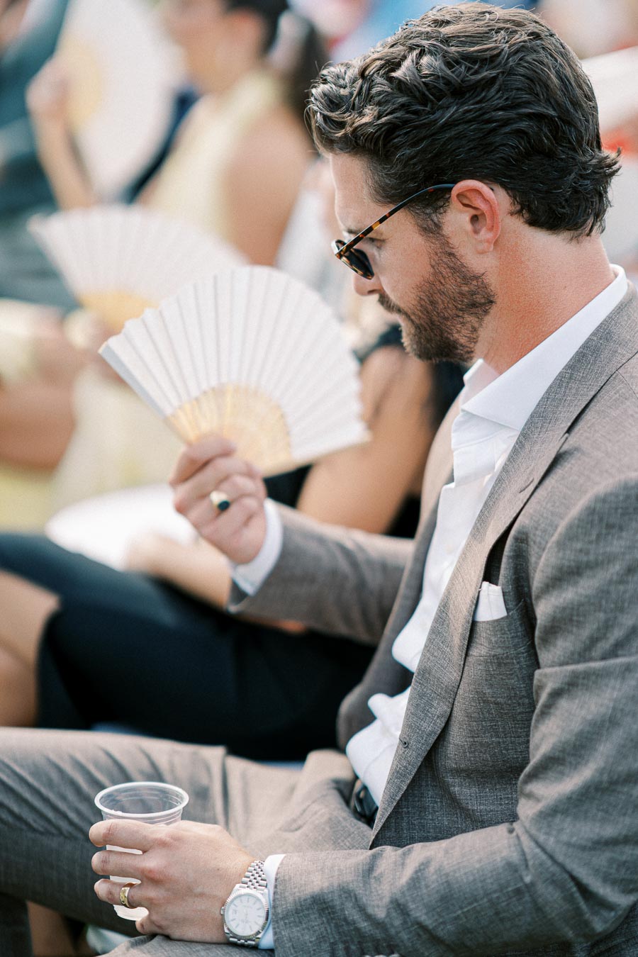 A man in a stylish gray suit and sunglasses sitting and holding a paper fan and a plastic cup.