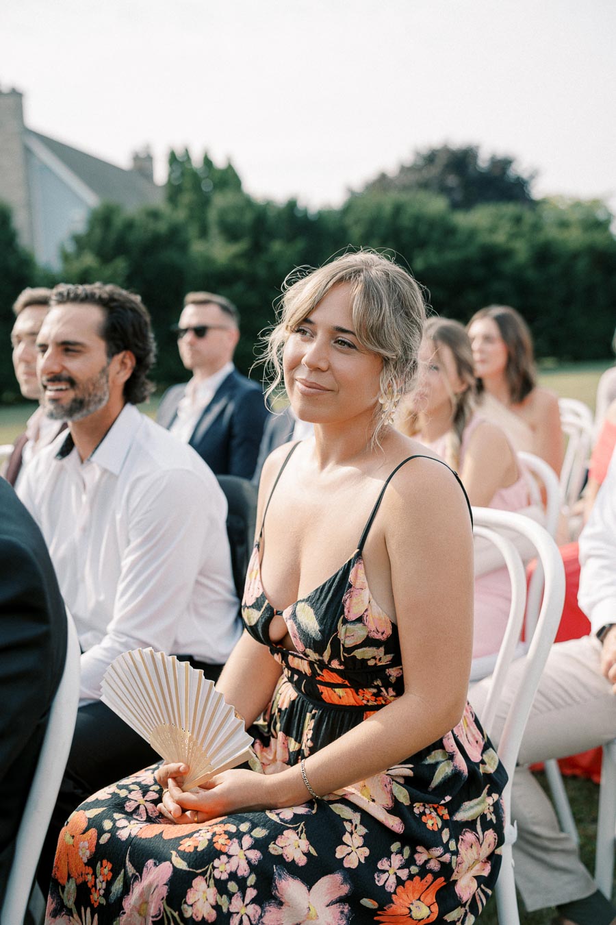 Woman in a floral dress holding a hand fan, seated outdoors at an event with other attendees in the background.