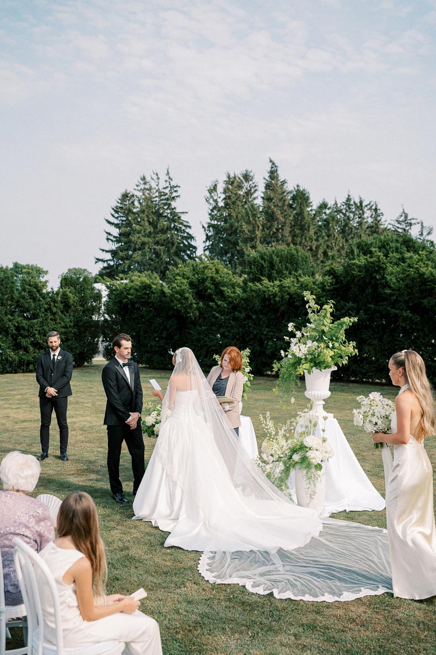 Elegant outdoor wedding ceremony with bride in white gown and long veil exchanging vows with groom in black tuxedo, surrounded by lush greenery and floral arrangements.