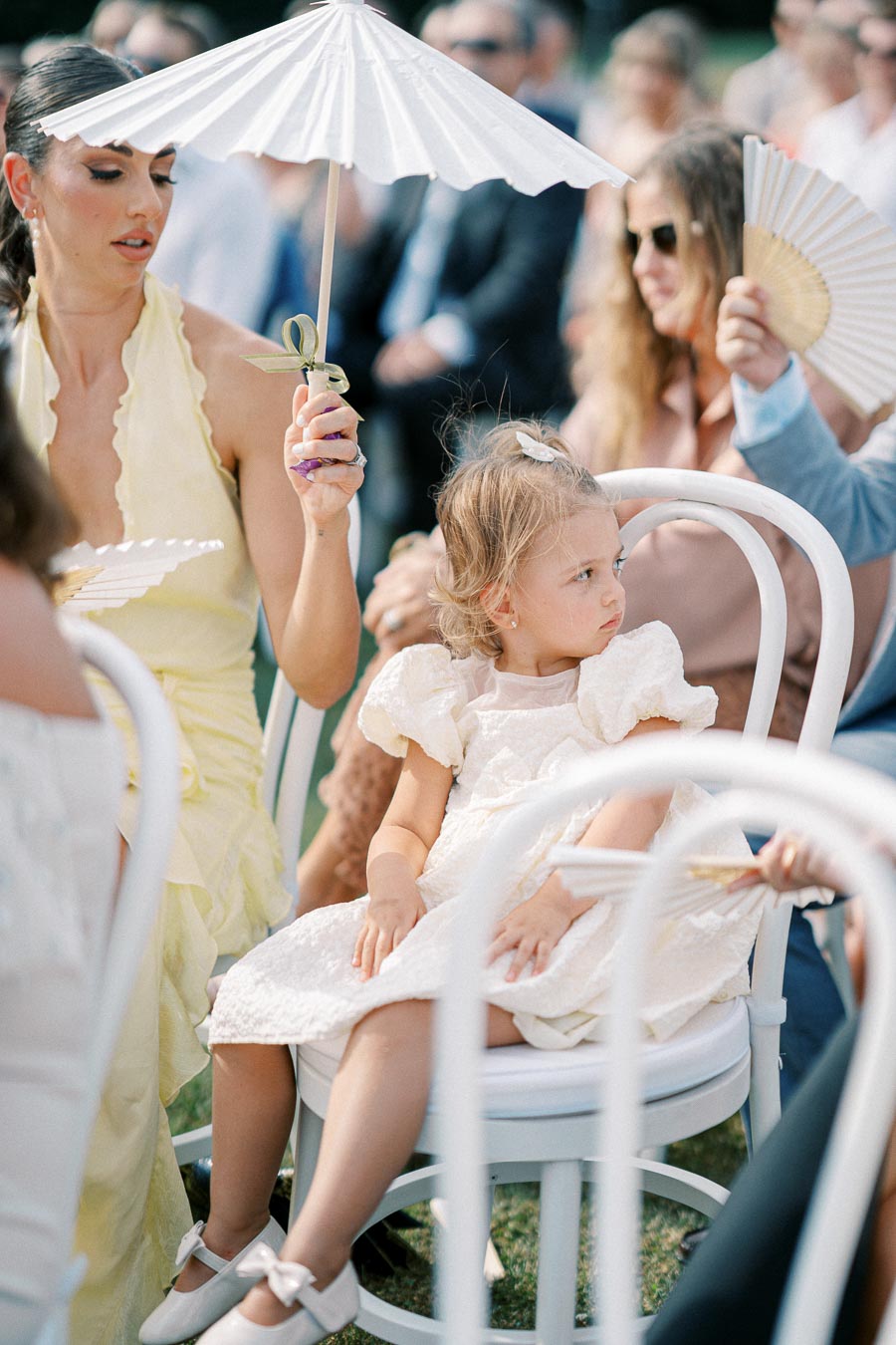 A young woman in a light yellow dress holds a white parasol while sitting next to a toddler in a frilly white dress at an outdoor event, surrounded by people holding fans. The focus is on the serene, sunlit setting.