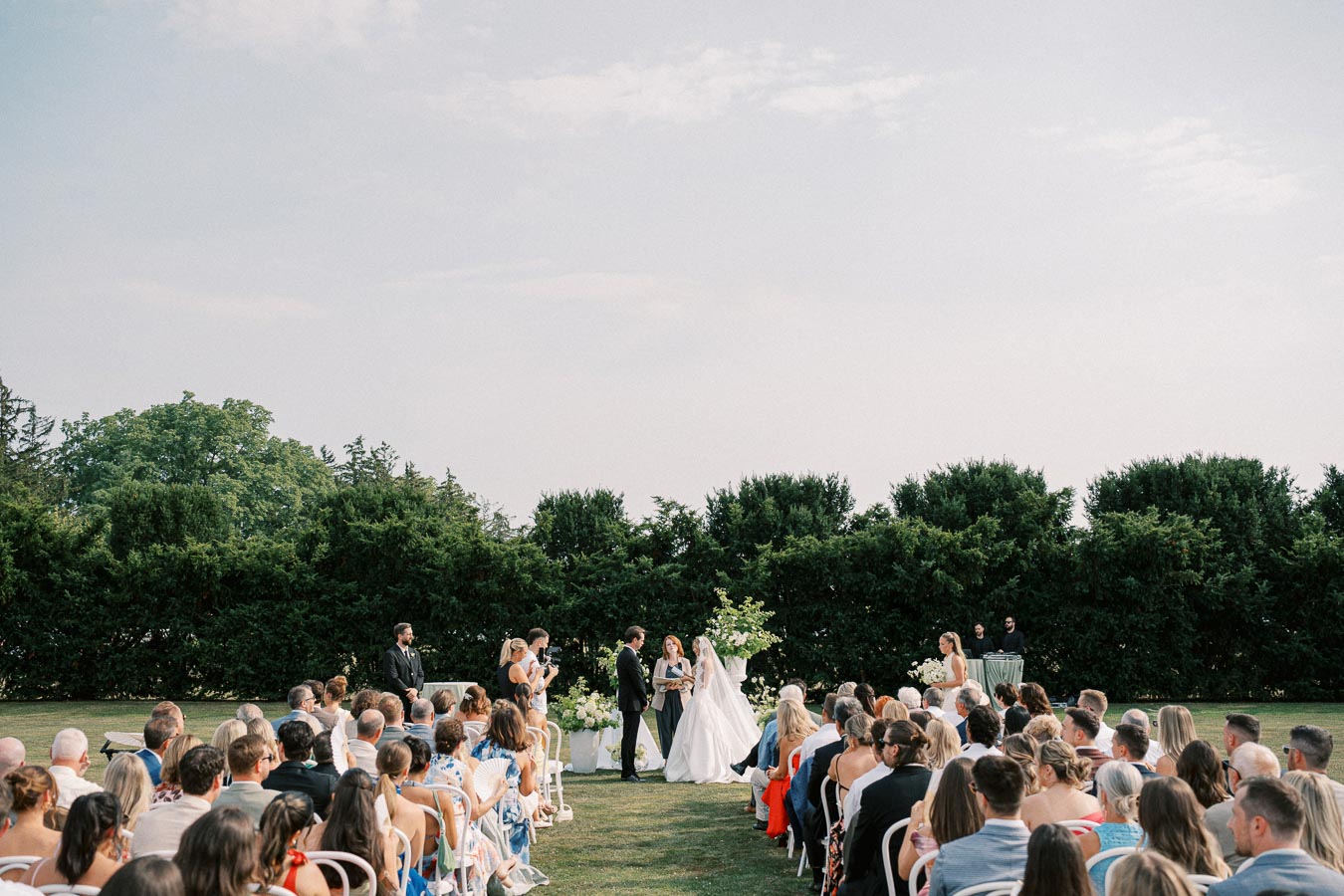 Outdoor wedding ceremony with a couple standing at the altar, surrounded by guests seated on a grassy lawn under a clear sky.