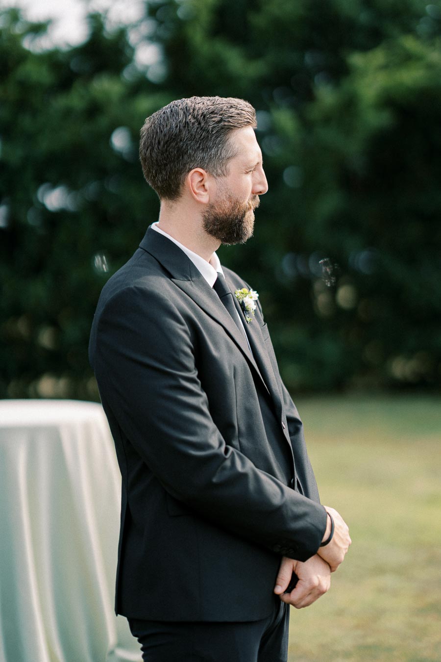 A man in a black suit stands outdoors with a thoughtful expression, surrounded by greenery.