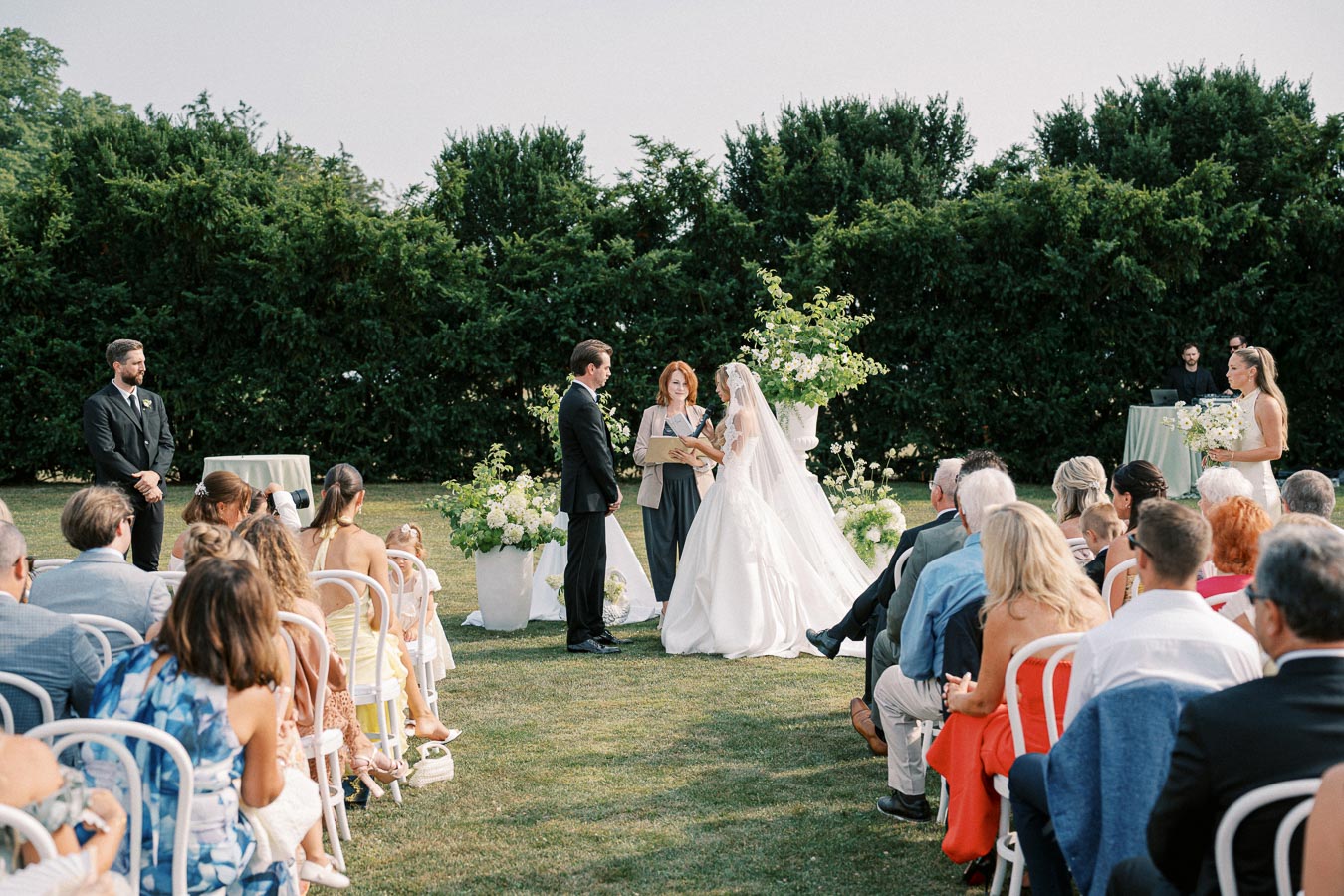 Outdoor wedding ceremony with a bride and groom exchanging vows in front of a celebrant. Guests are seated on white chairs, surrounded by greenery and floral arrangements under a clear sky.
