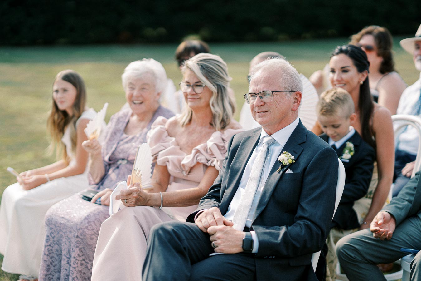 Group of wedding guests seated outdoors, including elderly and middle-aged individuals, dressed formally with some holding hand fans on a sunny day.