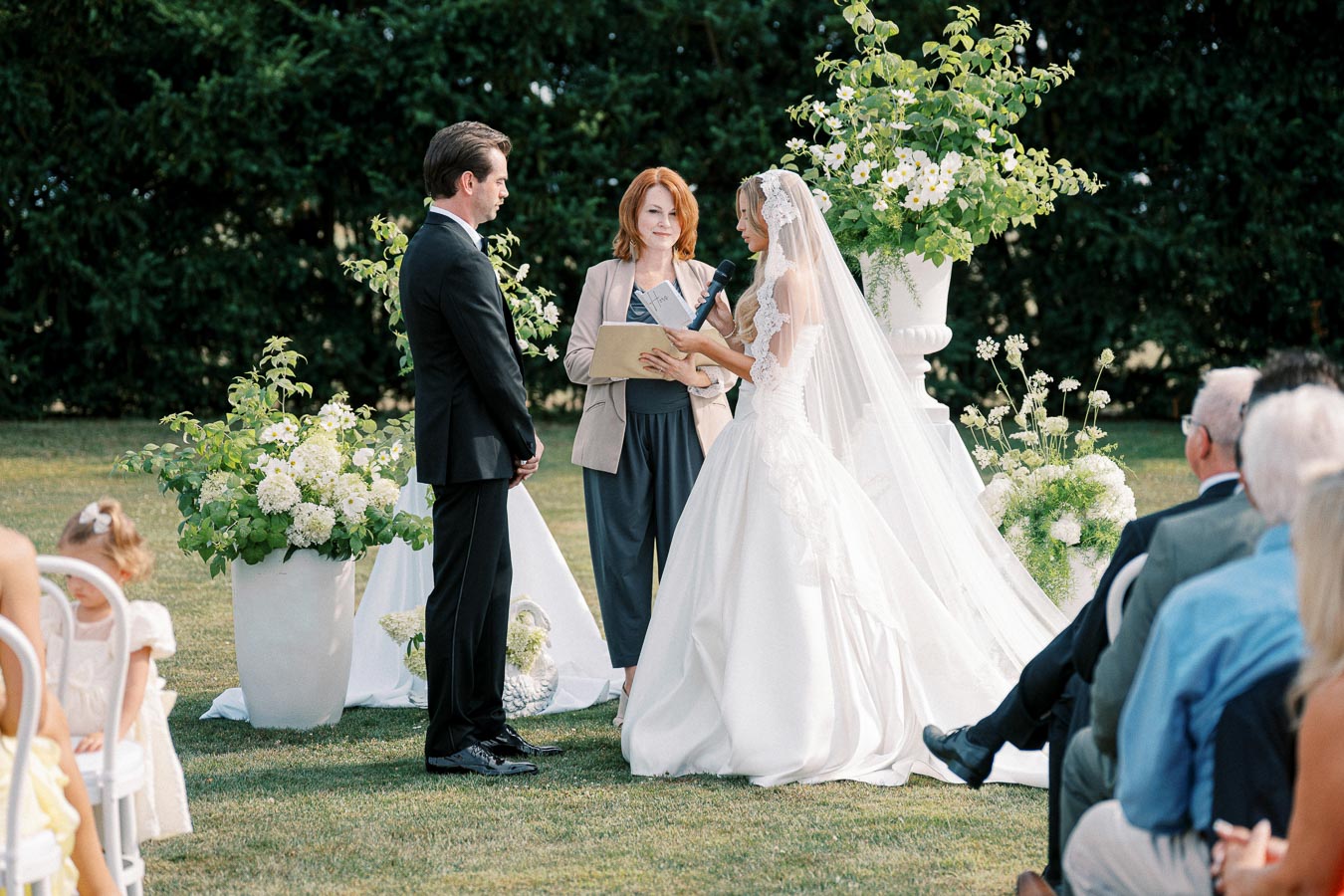 Outdoor wedding ceremony with a bride in a white gown and veil holding a microphone, standing next to a groom in a black suit, officiated by a red-haired woman. Surrounded by green foliage, white flowers, and seated guests on a sunny day.