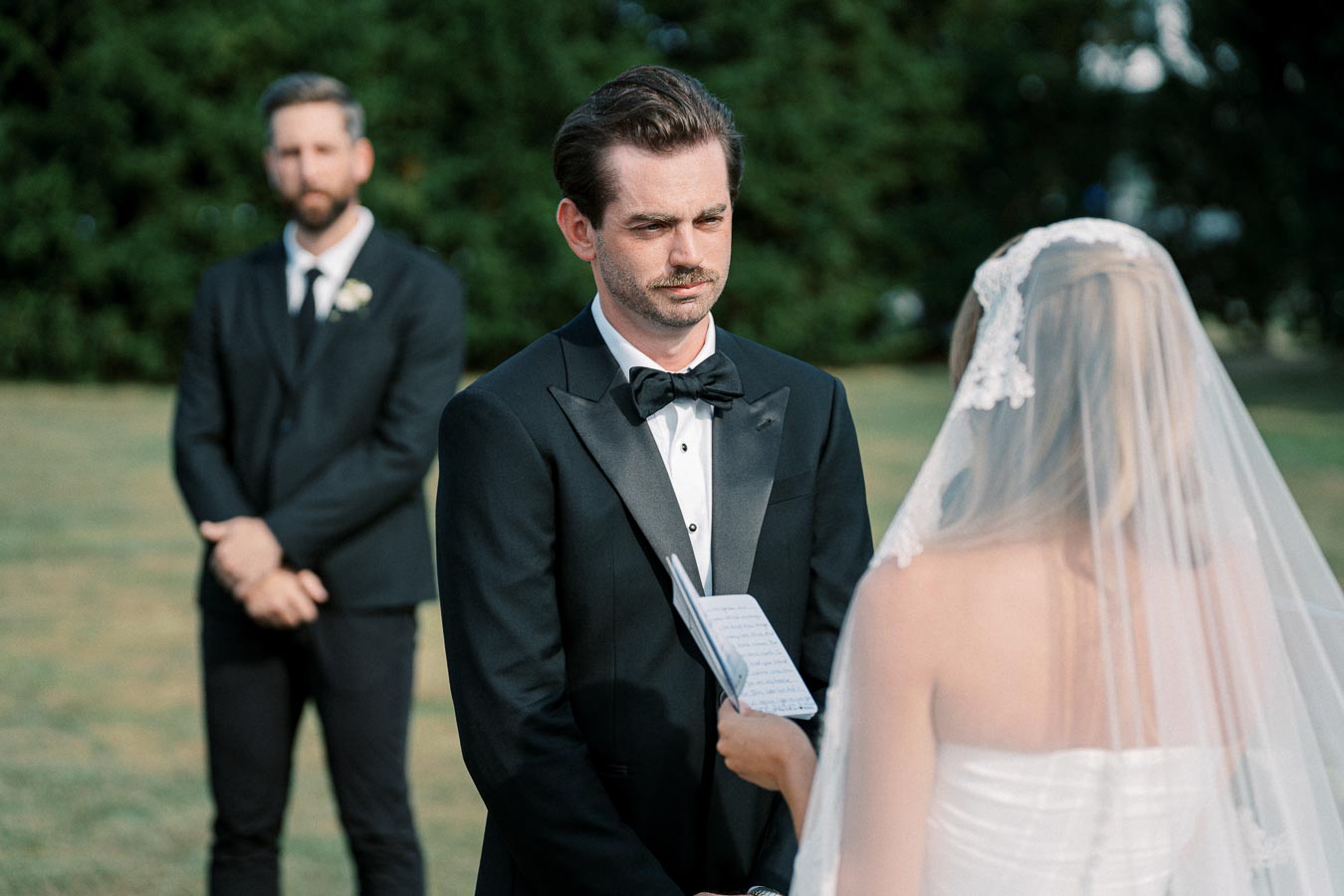 A bride in a white wedding dress holds a written note, facing a groom in a black tuxedo during an outdoor wedding ceremony, with a groomsman in the background, surrounded by greenery.
