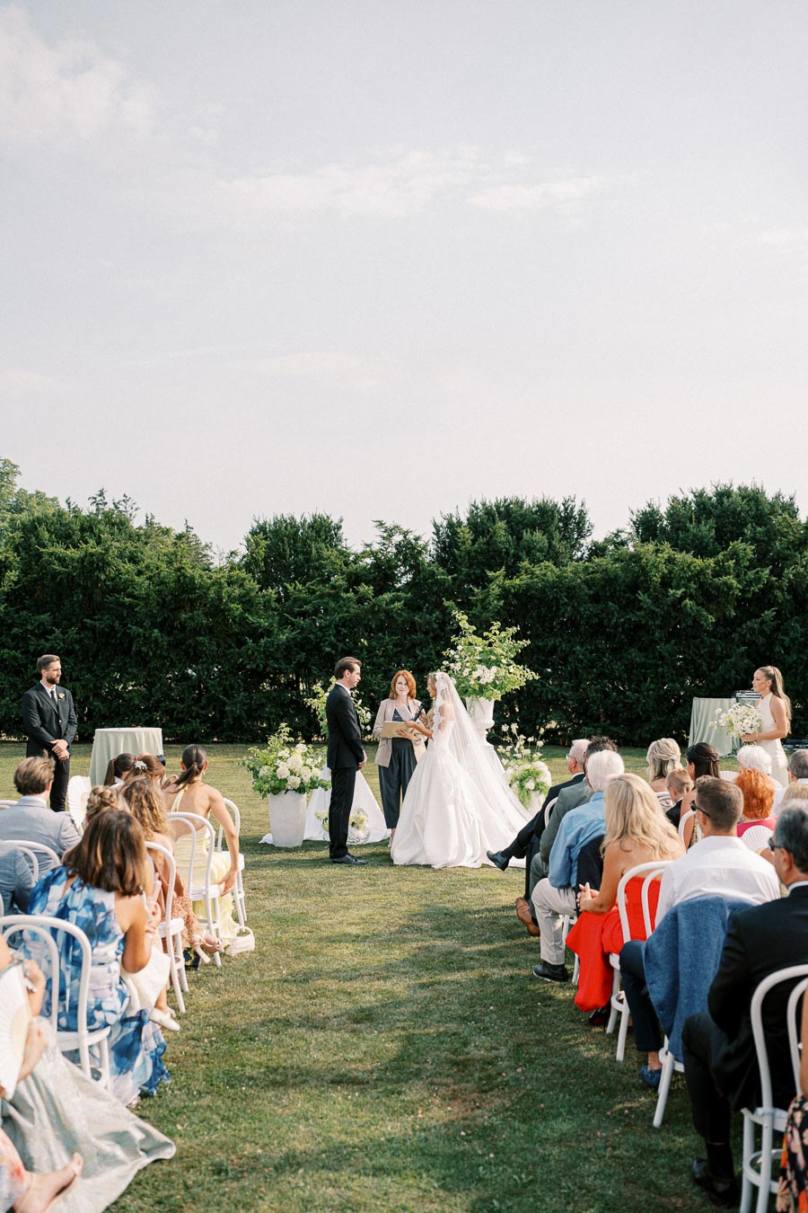 A picturesque outdoor wedding ceremony with guests seated on a lawn, a bride in a white gown and a groom in a black suit exchanging vows, surrounded by lush greenery and elegant floral arrangements under a clear sky.