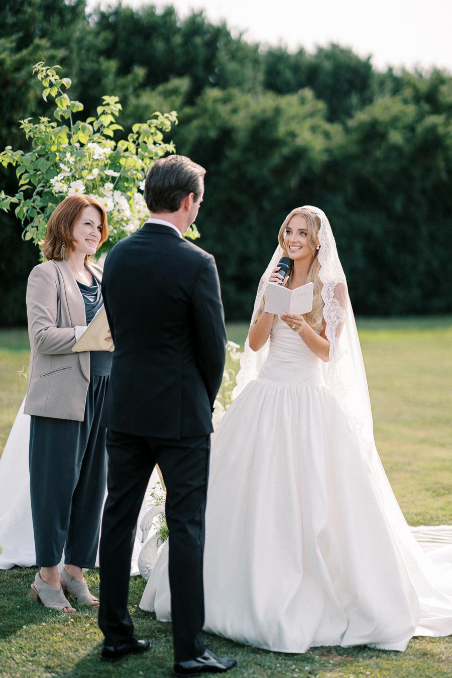 A bride in a white wedding dress reads vows to the groom during an outdoor ceremony, with an officiant standing nearby against a backdrop of greenery.