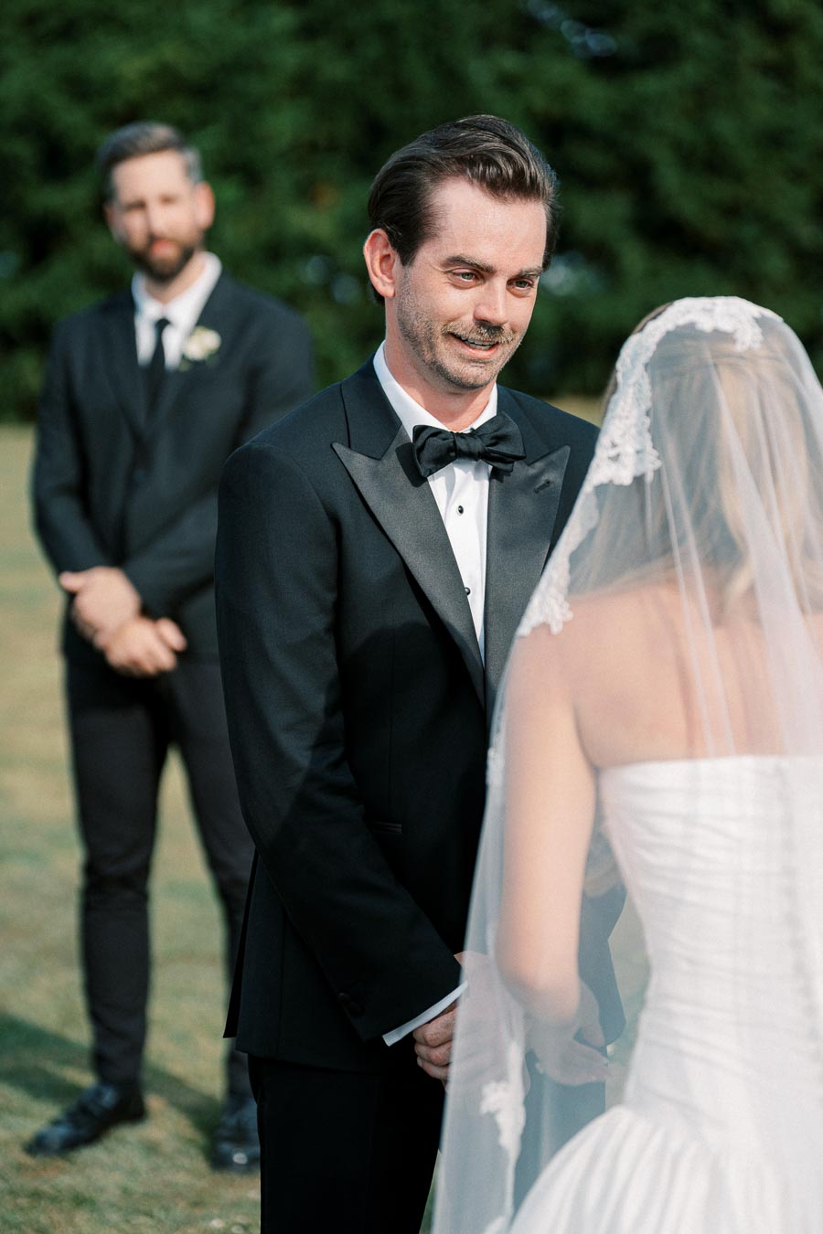 A groom in a black tuxedo smiles at his bride during an outdoor wedding ceremony, with a groomsman in the background.