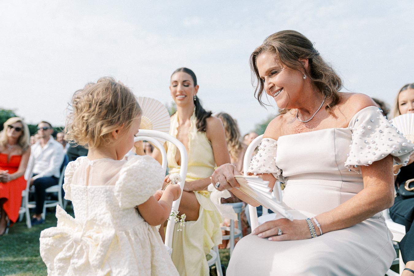 A joyful moment at an outdoor wedding ceremony, featuring a smiling woman in an elegant dress interacting with a young child wearing a white dress, surrounded by seated guests on a sunny day.