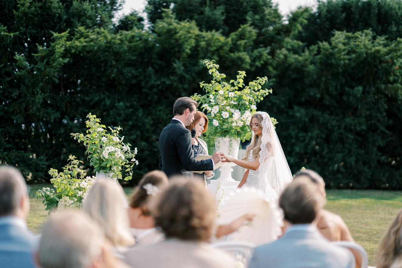 Outdoor wedding ceremony with a bride in a lace veil exchanging vows with a groom in a suit. A lush green backdrop and white floral arrangements enhance the romantic setting, attended by seated guests in festive attire.