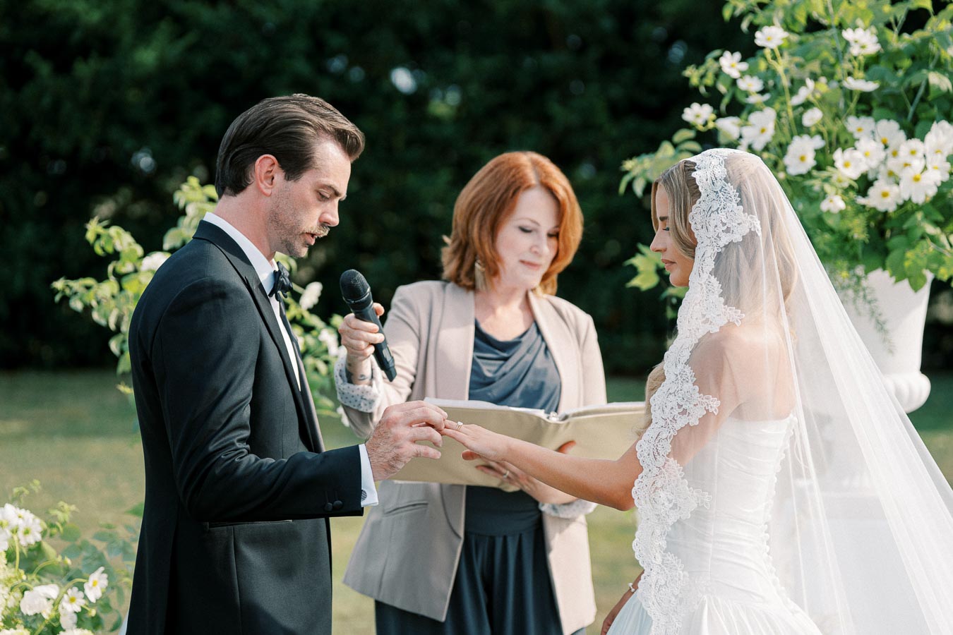 A bride and groom exchanging rings during an outdoor wedding ceremony, with an officiant holding a microphone and book, surrounded by floral decorations.