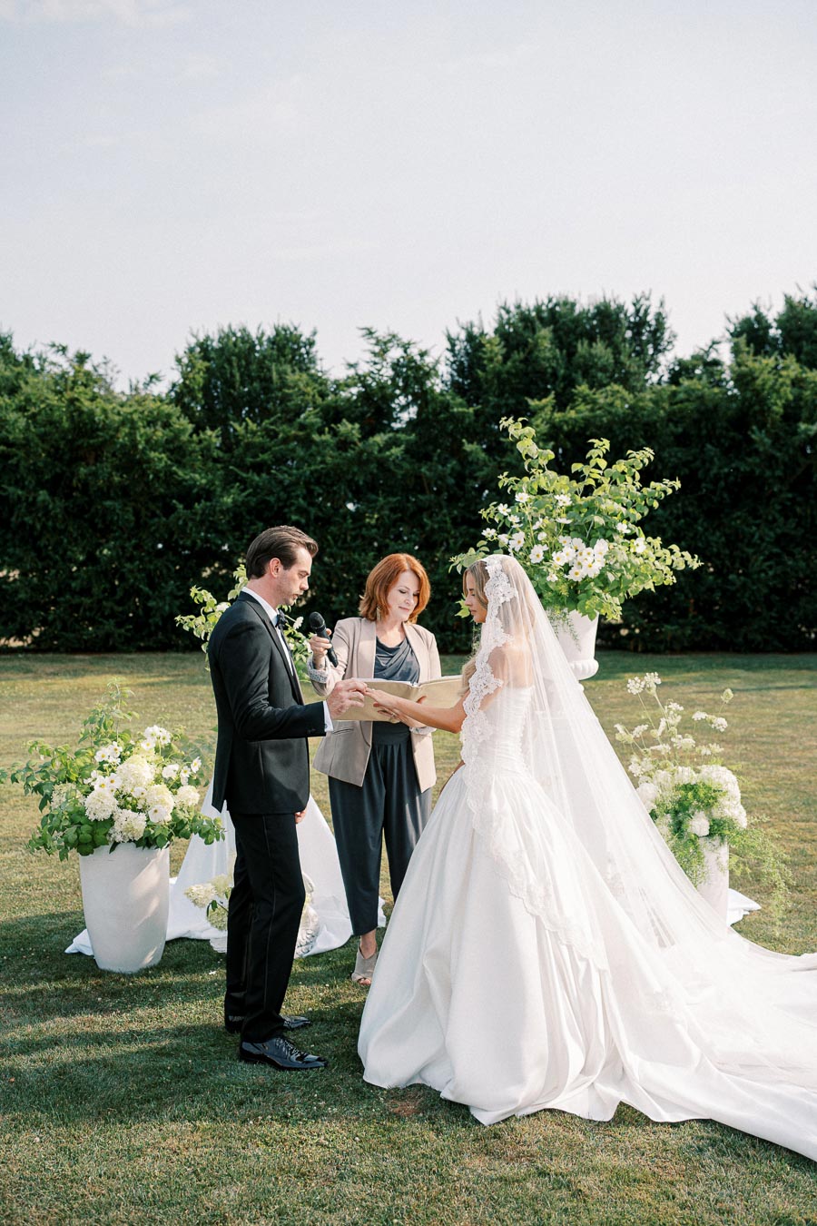 A bride and groom exchange vows during an outdoor wedding ceremony, with an officiant holding a microphone. The bride wears an elegant lace veil and gown, while the groom is in a black suit. They are surrounded by white floral arrangements and greenery on a sunny day.