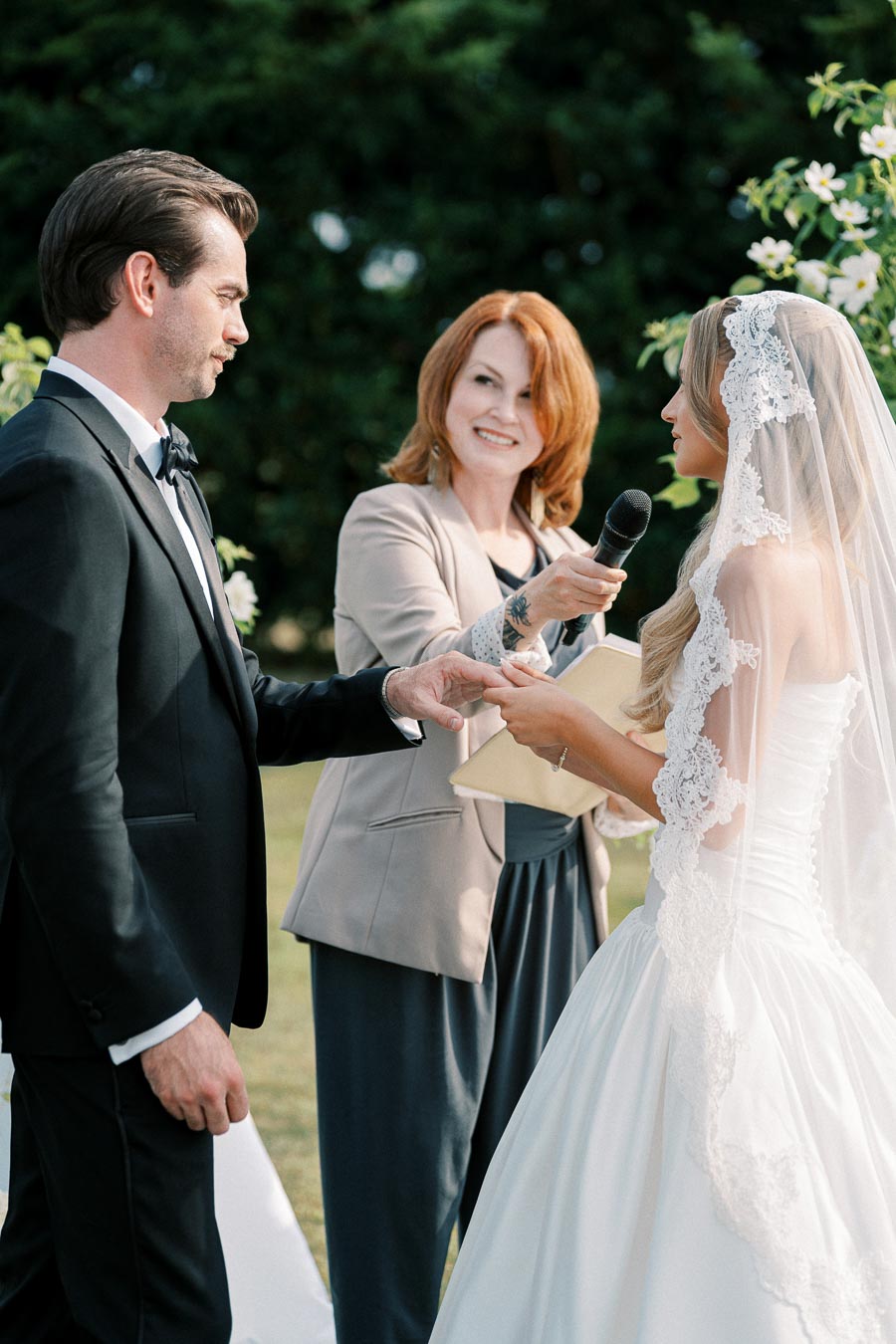 Bride and groom exchanging wedding vows outdoors, officiated by a woman holding a microphone.