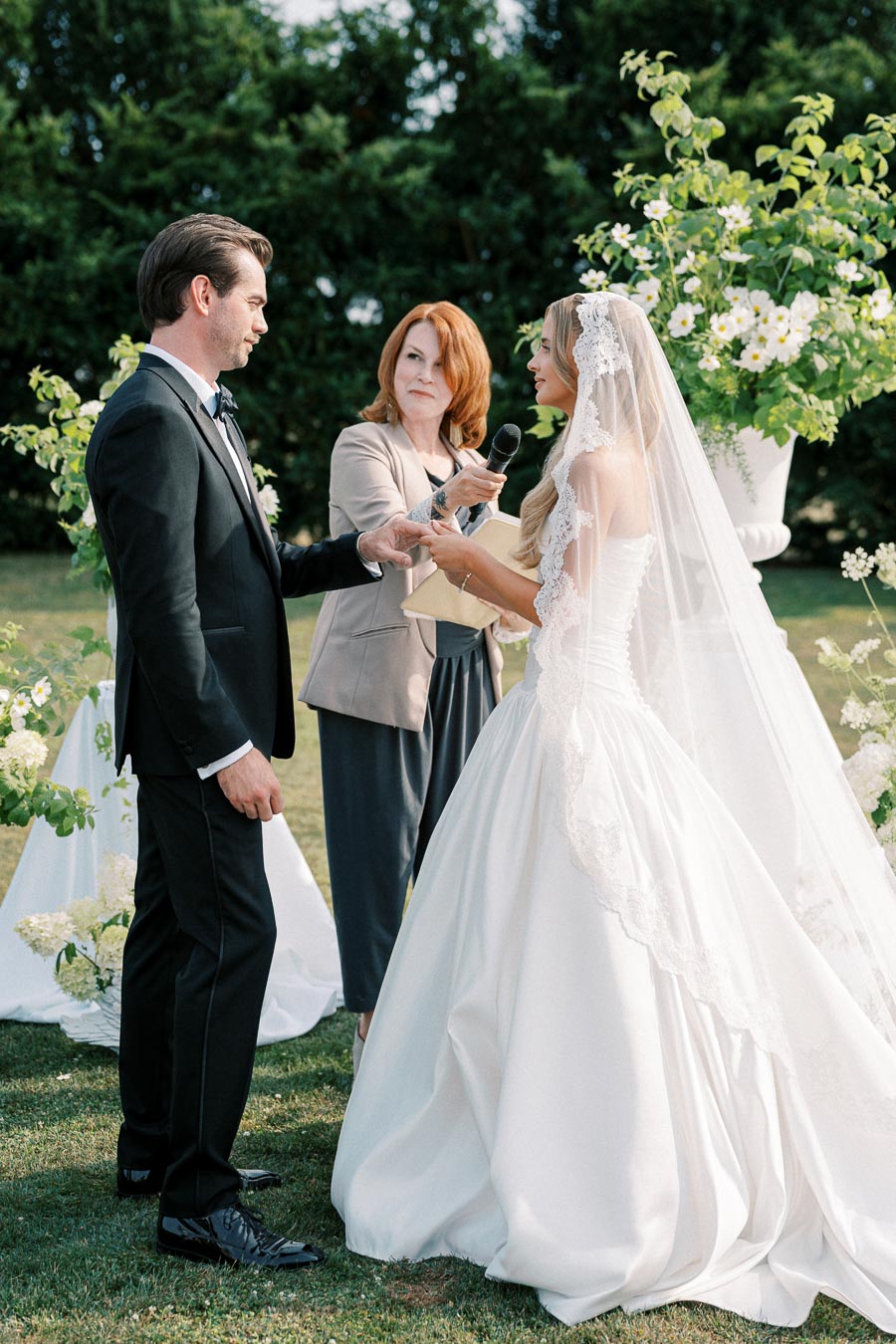 A bride and groom exchanging vows during an outdoor wedding ceremony, with an officiant holding a microphone, surrounded by green foliage and white flowers.