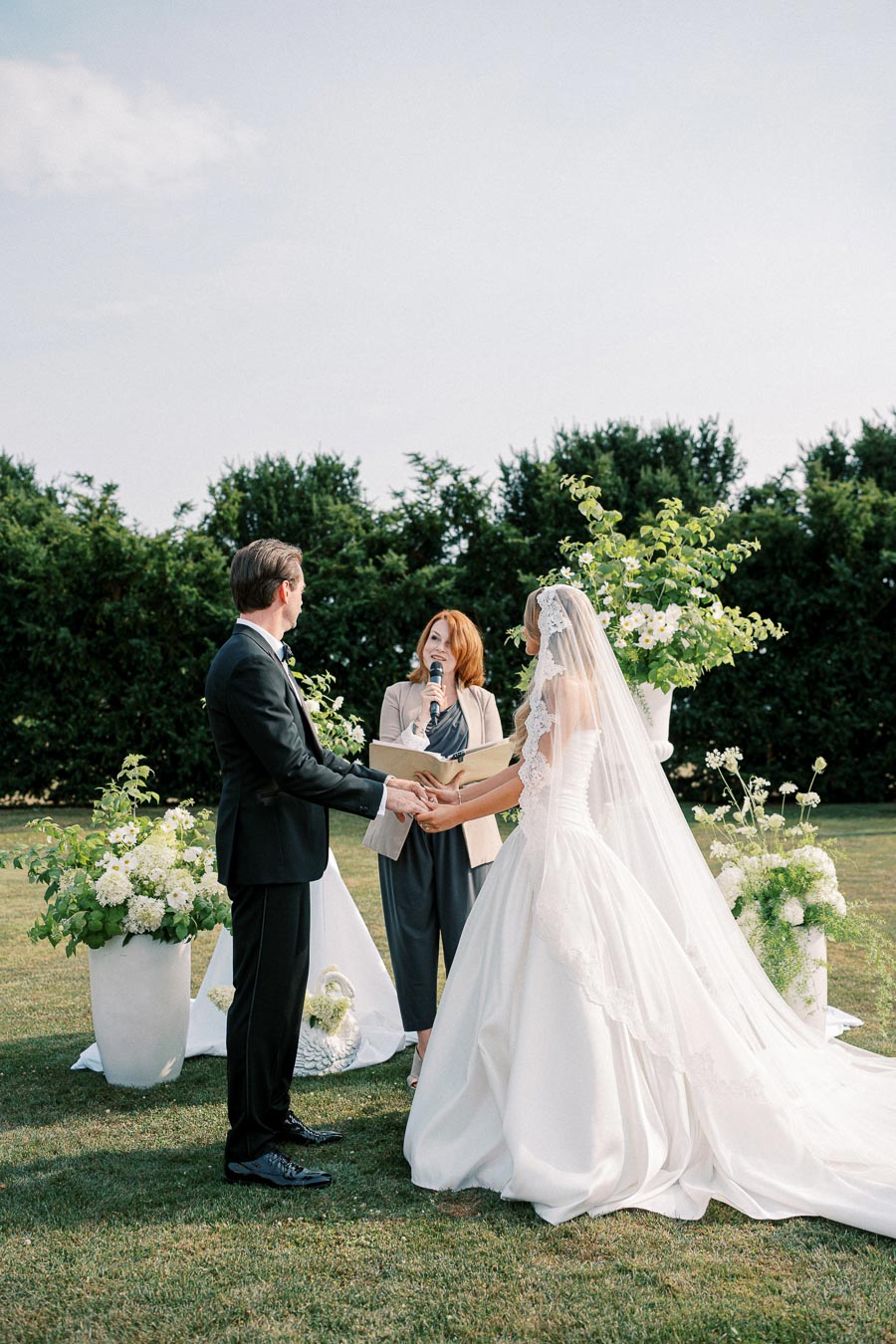 Outdoor wedding ceremony with bride in elegant white gown and groom in black suit holding hands, officiated by a woman with a microphone, surrounded by greenery and floral arrangements.