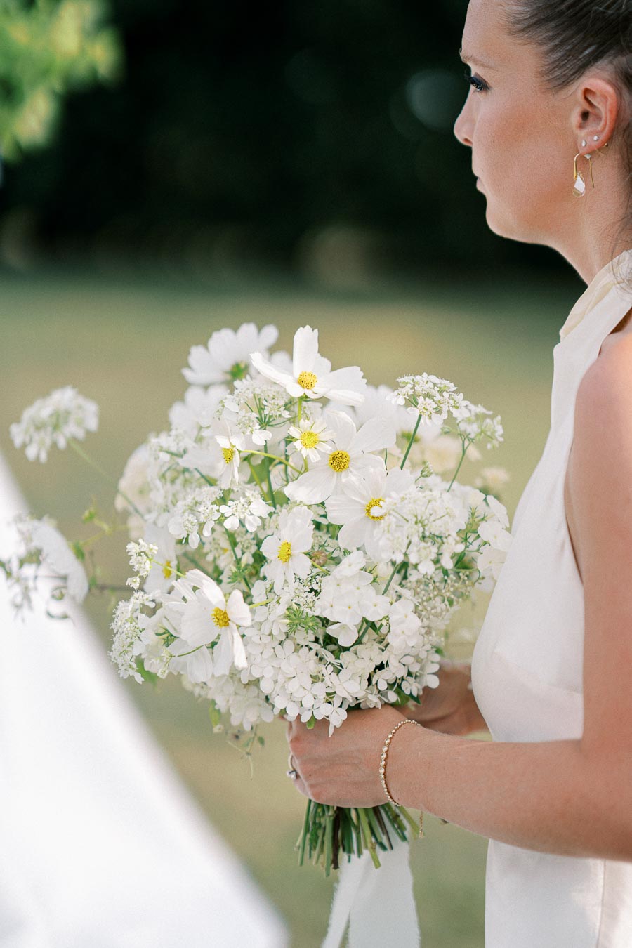 A woman in a sleeveless dress holds a bouquet of white flowers, including daisies and hydrangeas, in a garden setting.