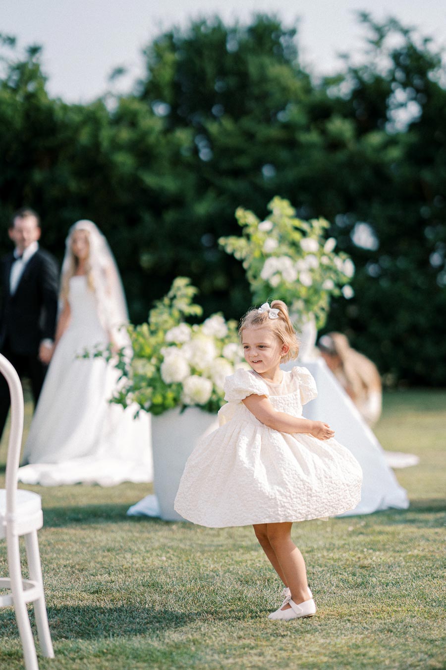 A young girl in a white dress twirling joyfully on a grassy lawn at an outdoor wedding, with a blurred background of a bride and groom standing in front of lush greenery and floral arrangements.