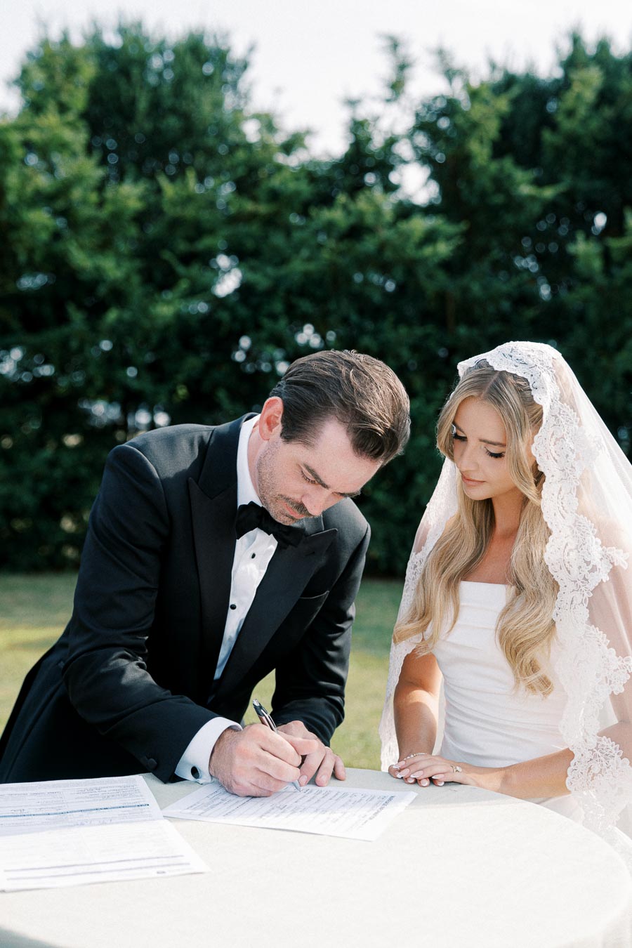 A groom in a black tuxedo signs a wedding document while the bride, in a white dress and lace veil, looks on during an outdoor ceremony with greenery in the background.