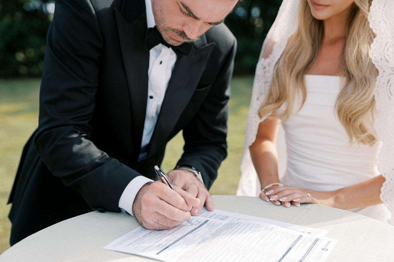 A groom in a black tuxedo signing a marriage certificate on a table, with a bride in a white dress standing beside him during an outdoor wedding ceremony.