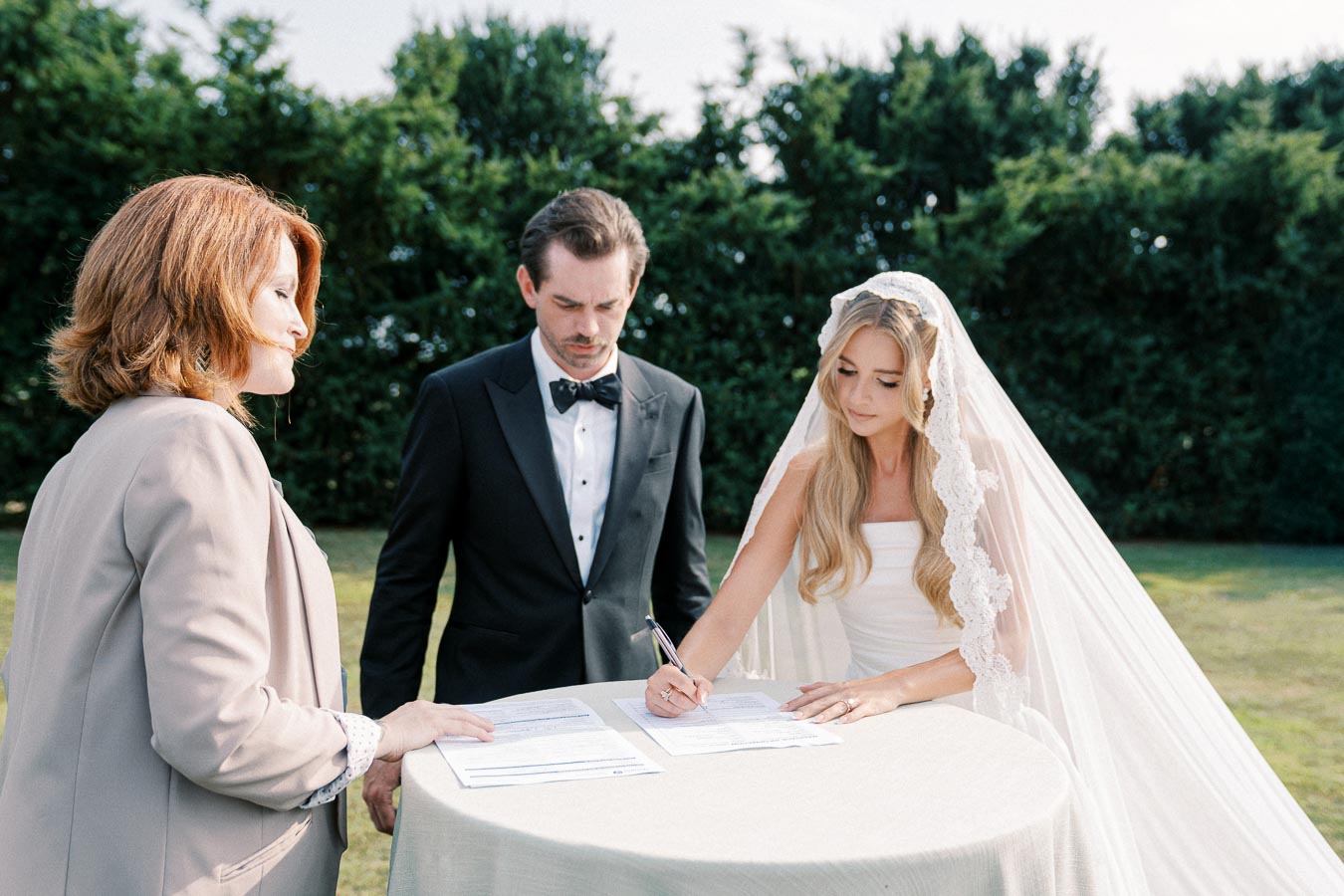 Bride signing wedding certificate outdoors with groom and officiant nearby.