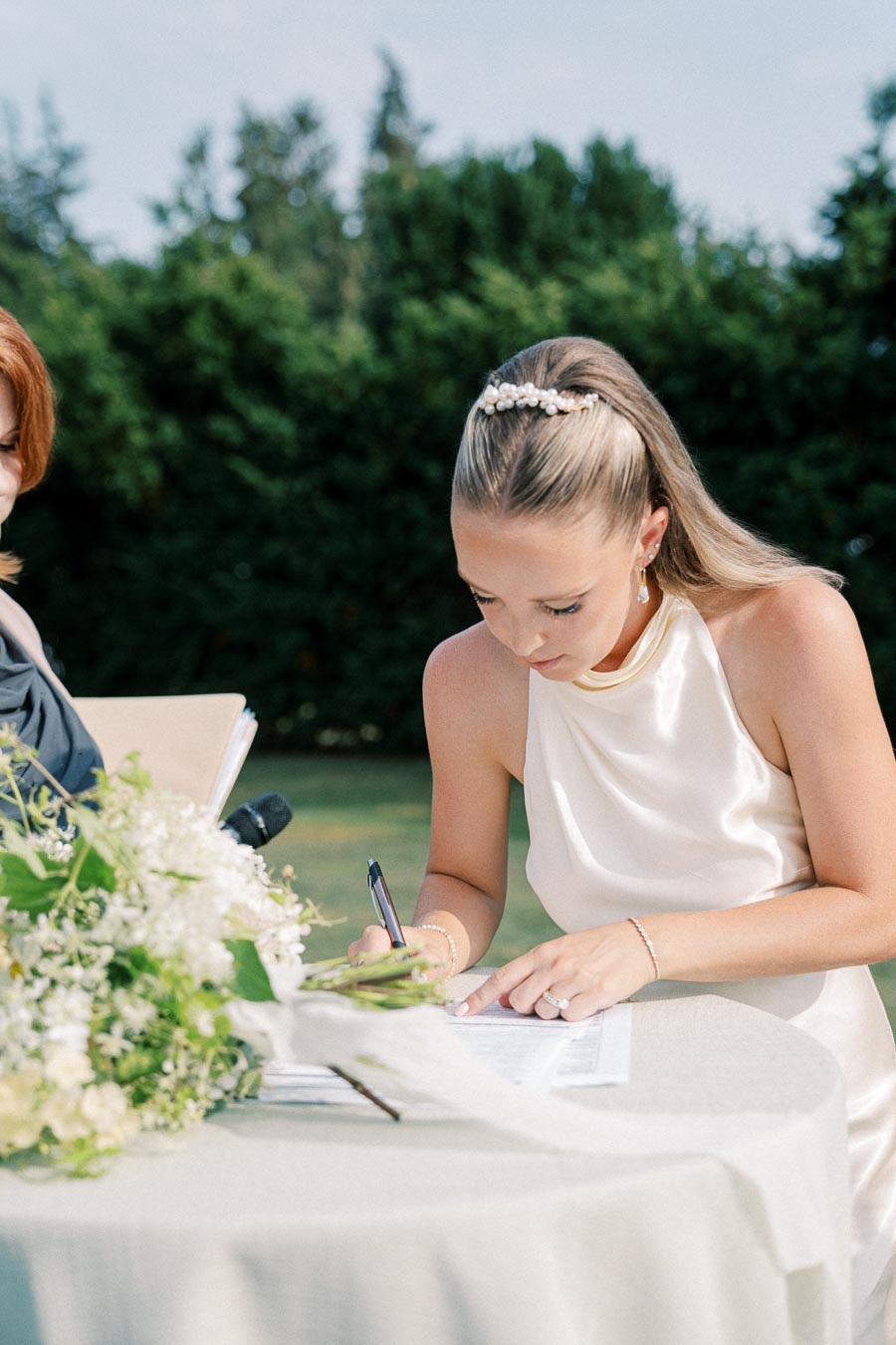 A bride in a white satin dress signs a document at an outdoor wedding ceremony, with a bouquet of flowers on the table and greenery in the background.