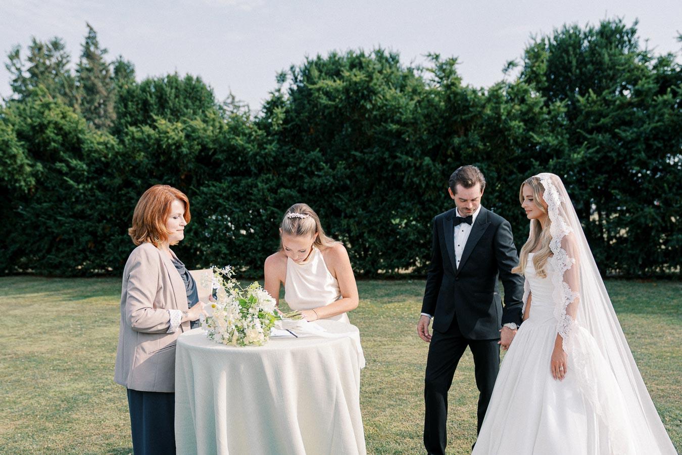 Bride and groom signing marriage documents in outdoor wedding ceremony, accompanied by a woman in a suit, against a backdrop of lush greenery.