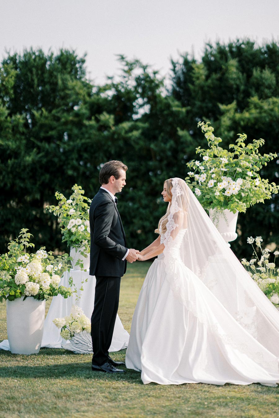 Elegant outdoor wedding ceremony with bride and groom holding hands, surrounded by floral arrangements, lush greenery backdrop, and a serene garden setting.
