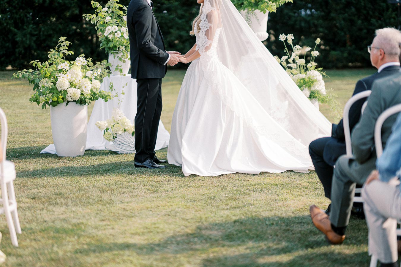 A bride and groom holding hands during an outdoor wedding ceremony, surrounded by lush greenery and floral arrangements, with seated guests in the foreground.