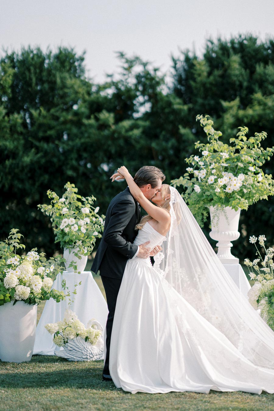 Elegant outdoor wedding ceremony with a bride in a flowing white gown and veil kissing the groom in a black tuxedo, surrounded by lush greenery and white floral arrangements.