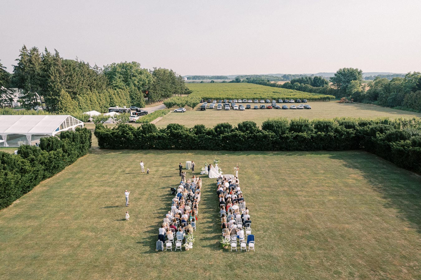Aerial view of an outdoor wedding ceremony in a lush green field, with guests seated on white chairs in two rows facing a wedding arch. The background features parked cars and expansive greenery, providing a picturesque rural setting.