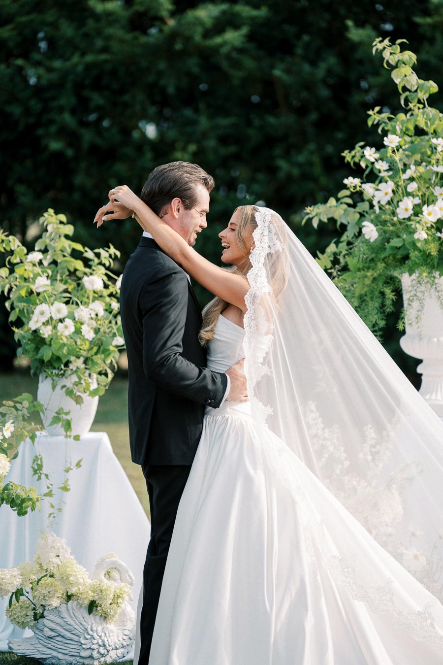 A bride and groom embrace during an outdoor wedding ceremony, surrounded by lush greenery and elegant white floral arrangements. The bride wears a flowing white gown with a lace veil, while the groom is dressed in a dark suit.