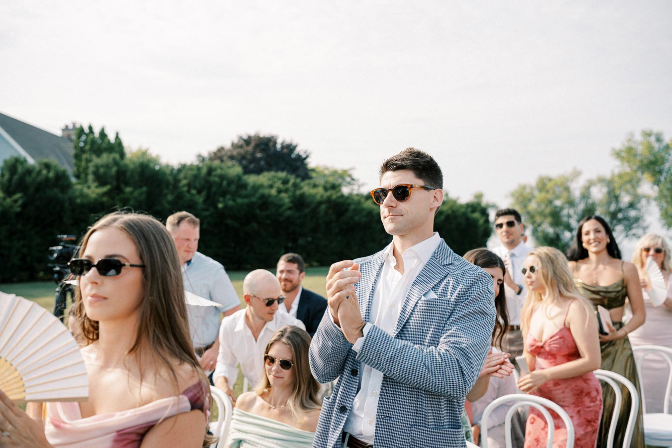 Group of elegantly dressed wedding guests clapping at an outdoor ceremony, with a man in a checkered blazer and sunglasses standing prominently in the foreground, surrounded by greenery and a clear sky.