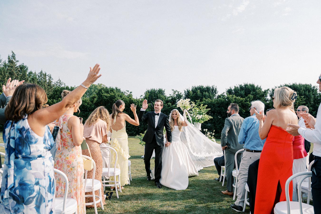 A newlywed couple joyfully walking down the aisle at an outdoor wedding ceremony, surrounded by cheering guests in colorful attire, with lush greenery in the background.