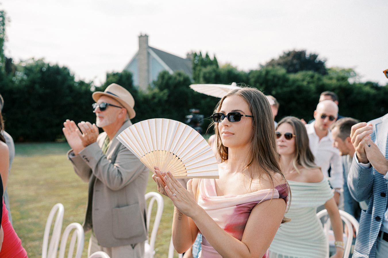 A stylish woman in sunglasses holds a hand fan at an outdoor event. She is surrounded by a diverse group of people clapping and enjoying a sunny day.