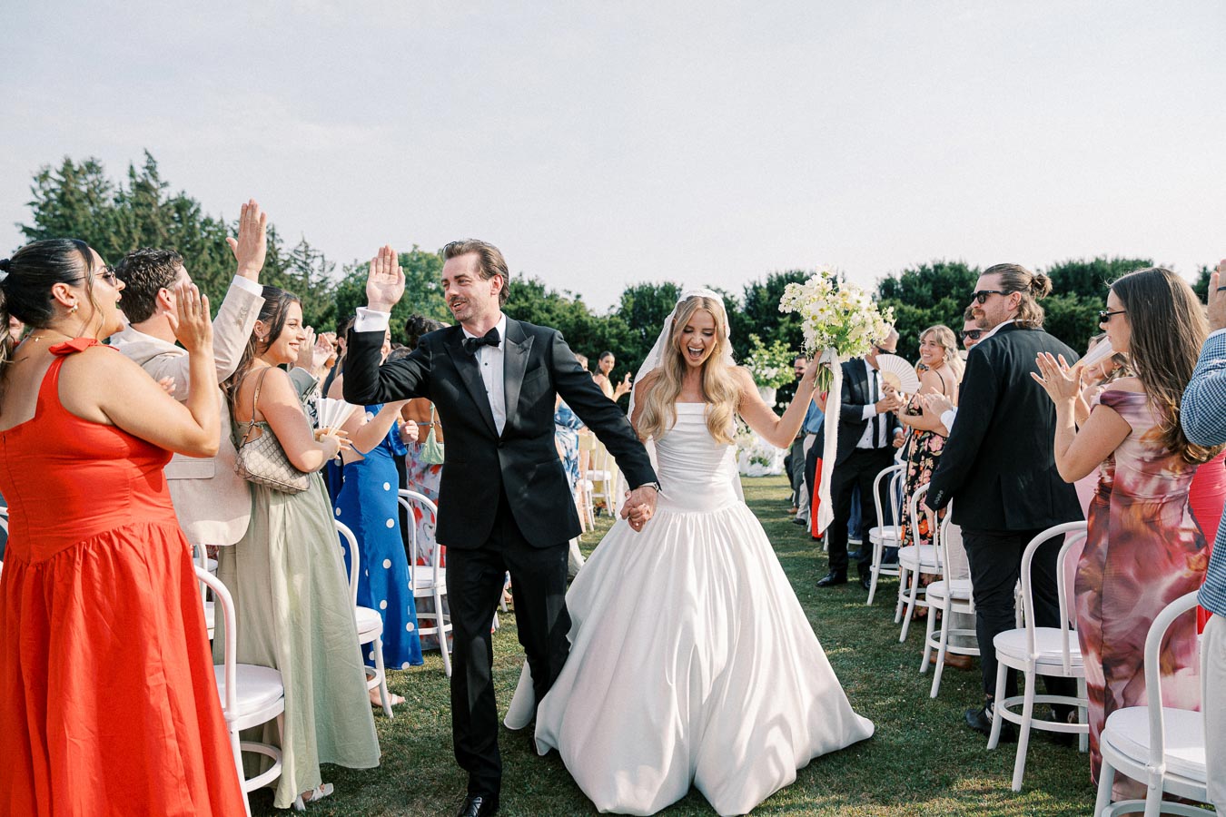 Bride and groom joyfully walking down the aisle, surrounded by applauding guests in colorful attire during an outdoor wedding ceremony.