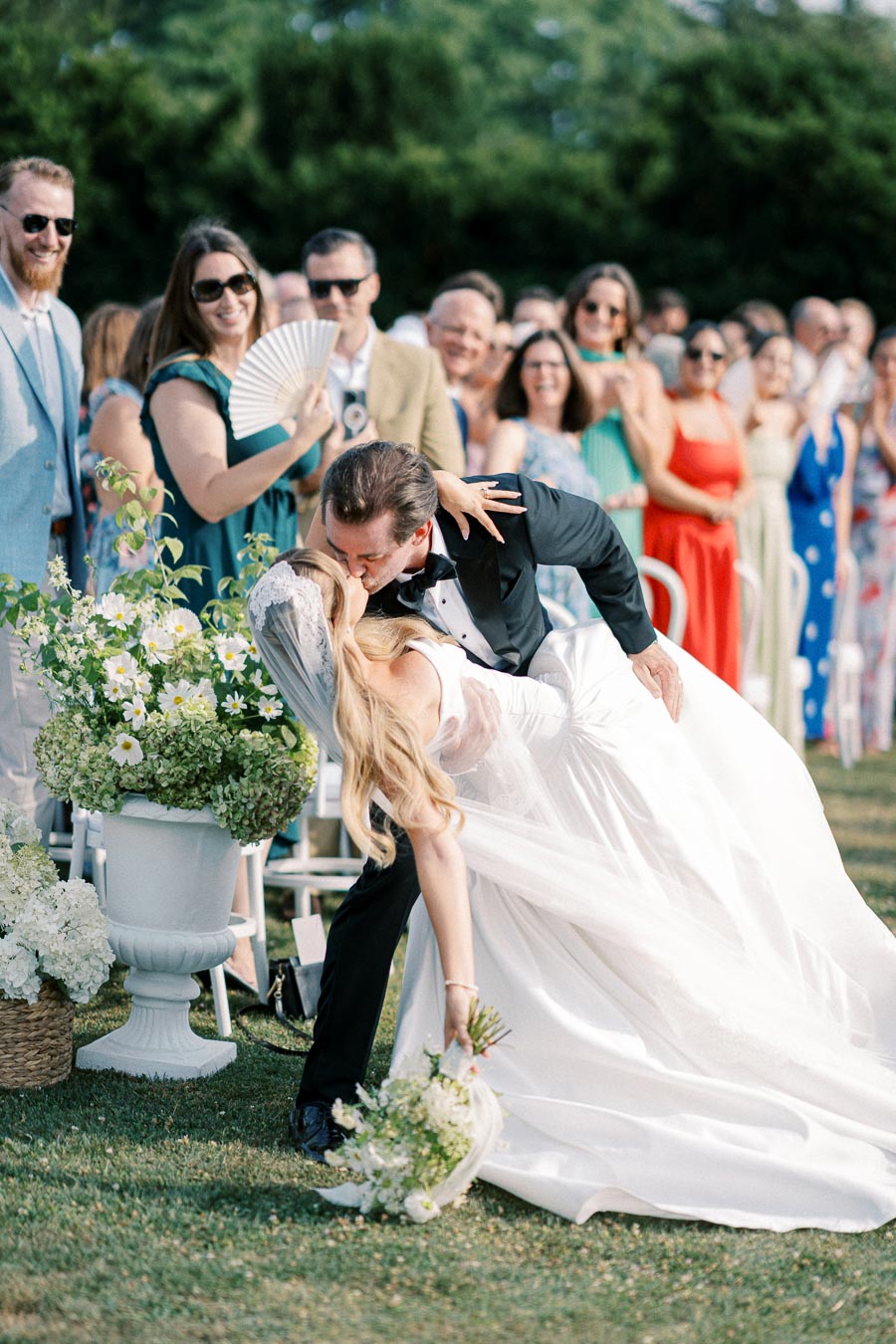 Bride and groom sharing a romantic kiss at an outdoor wedding ceremony, surrounded by celebrating guests and lush floral arrangements.