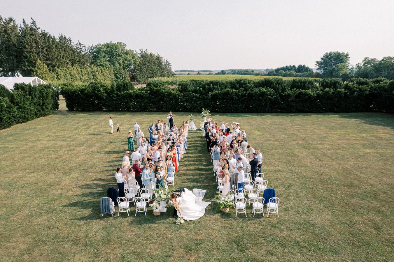 Aerial view of an outdoor wedding ceremony with guests seated on a lush green lawn. The couple is walking down the aisle, surrounded by neatly arranged chairs and scenic greenery.