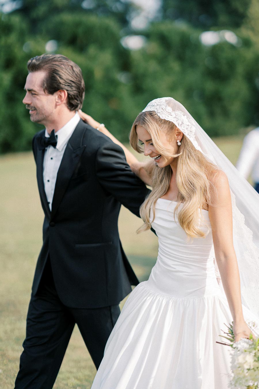 Smiling bride and groom walking outdoors in elegant wedding attire, with bride in a white gown and veil, and groom in a black tuxedo.