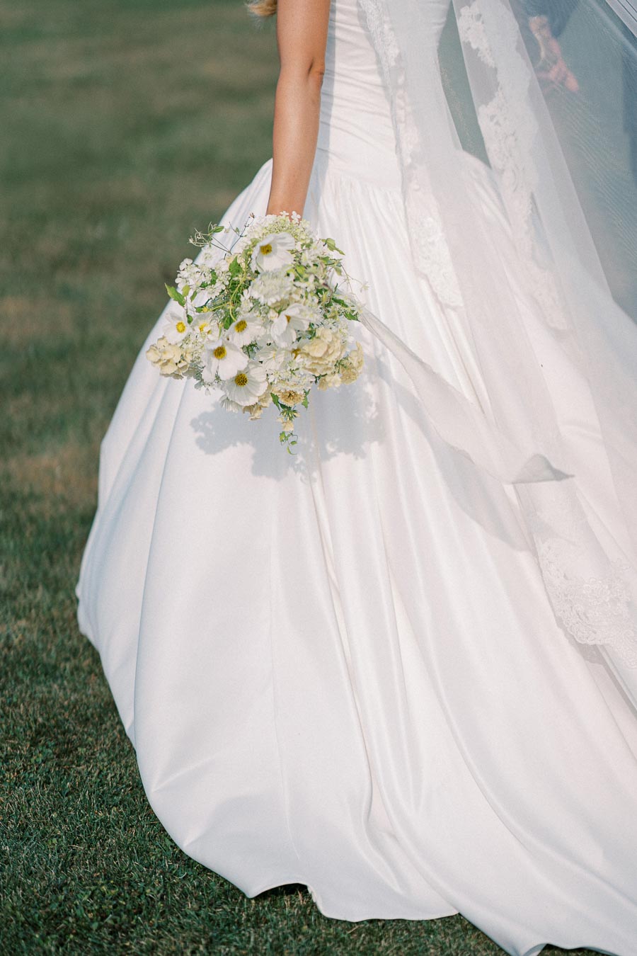 Bridal gown and floral bouquet close-up on grass background, highlighting elegant white wedding dress details and delicate bouquet of white and yellow flowers.