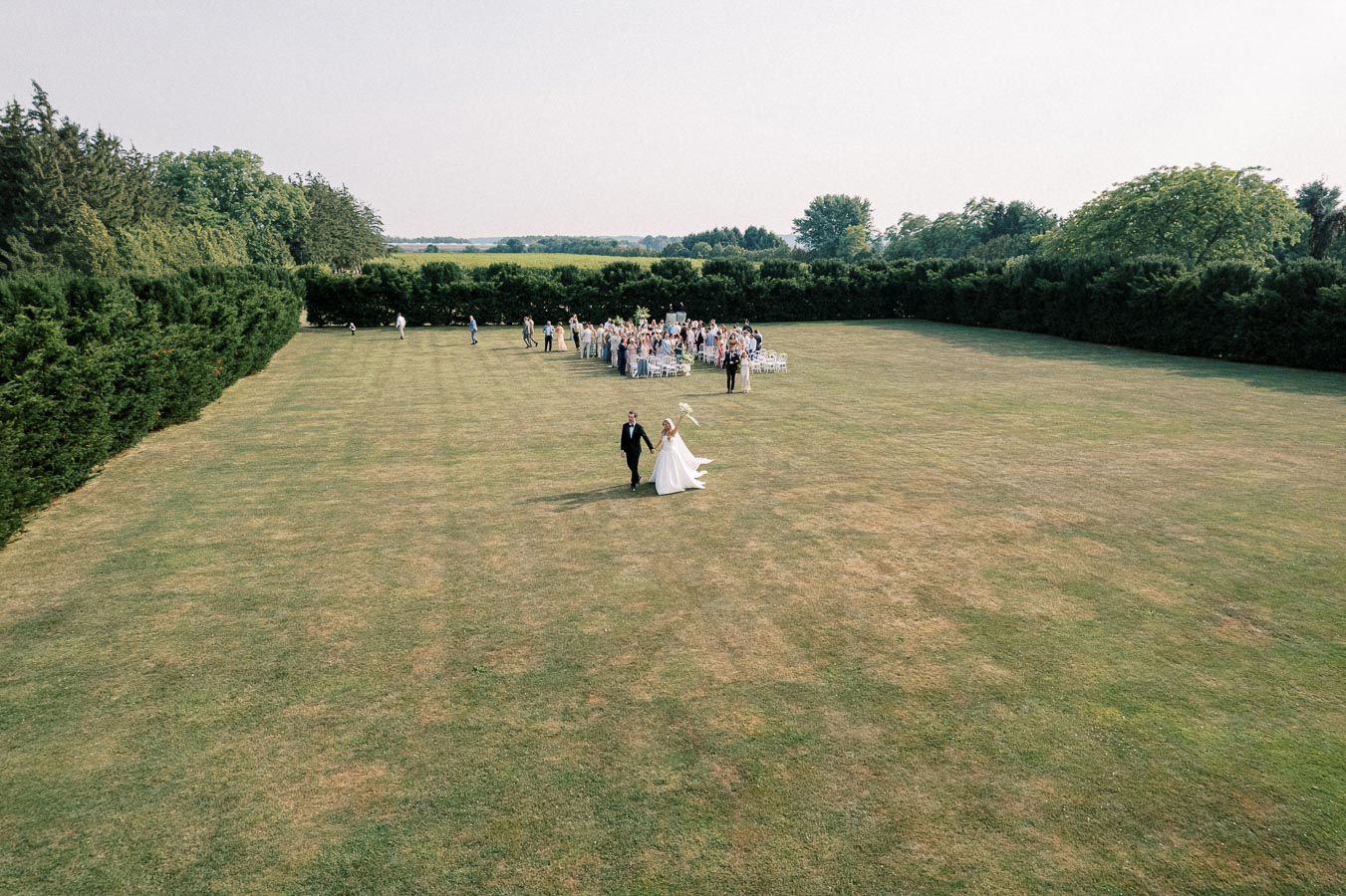 A newlywed couple walks hand-in-hand across a spacious, green lawn toward a large group of guests gathered for an outdoor wedding ceremony surrounded by lush trees.