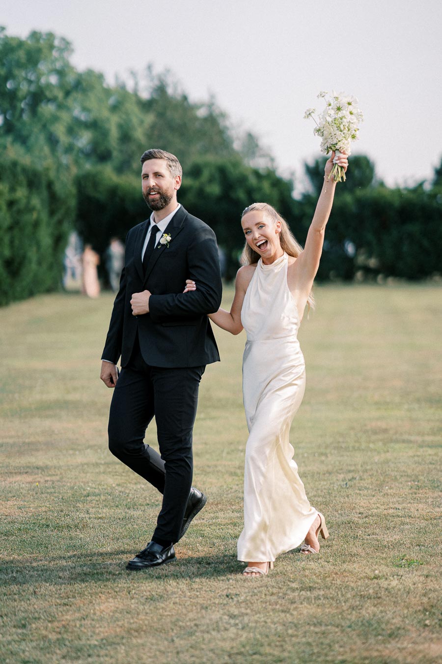 A joyful bride and groom walking arm in arm on grassy lawn, with the bride holding a bouquet of flowers triumphantly in the air.