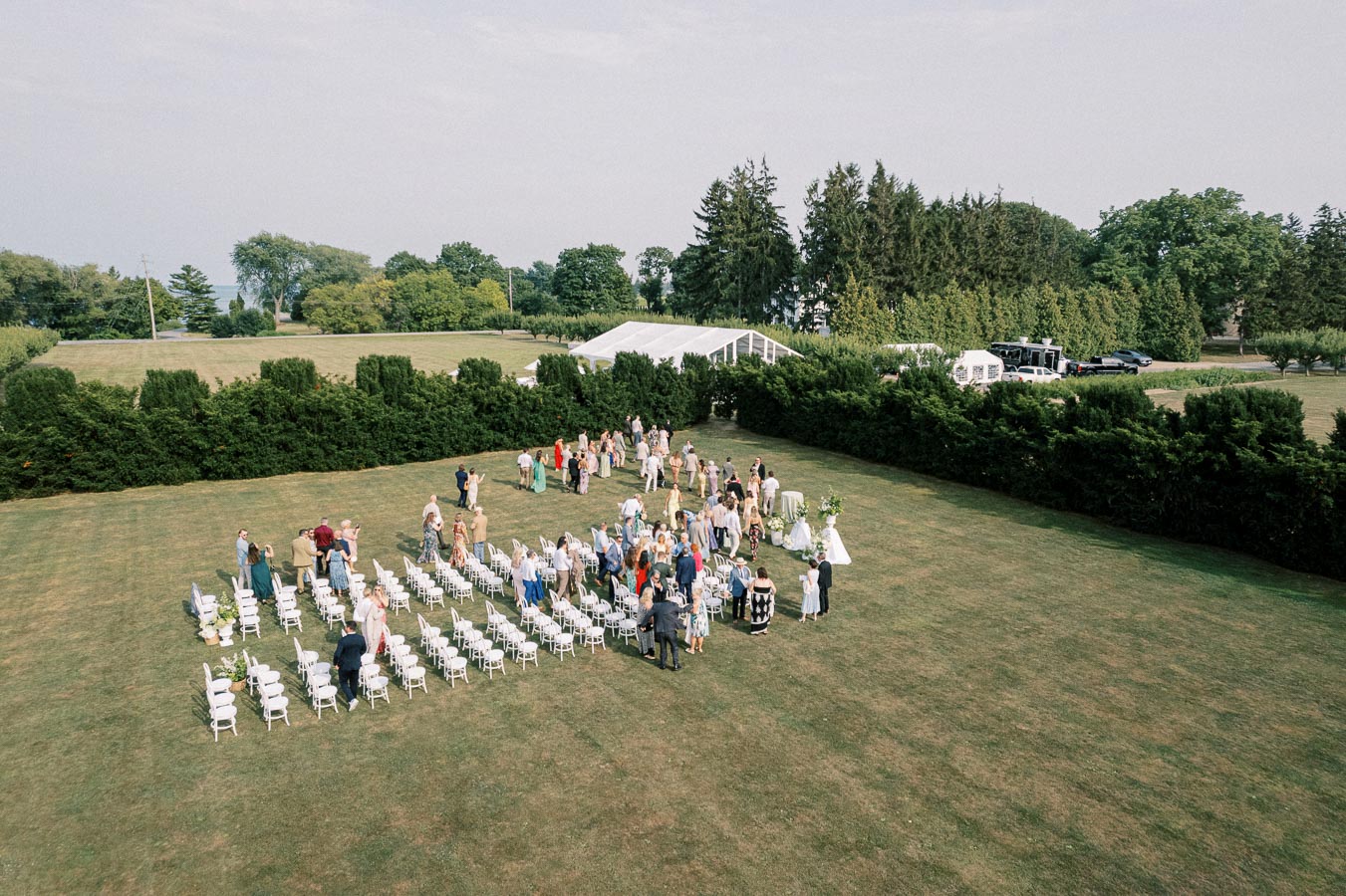 Aerial view of an outdoor wedding ceremony set up in a large, green field surrounded by trees. Rows of white chairs are arranged neatly, with guests gathering and socializing. A white tent is visible in the background, suggesting a reception area. The scene conveys a festive and elegant atmosphere.