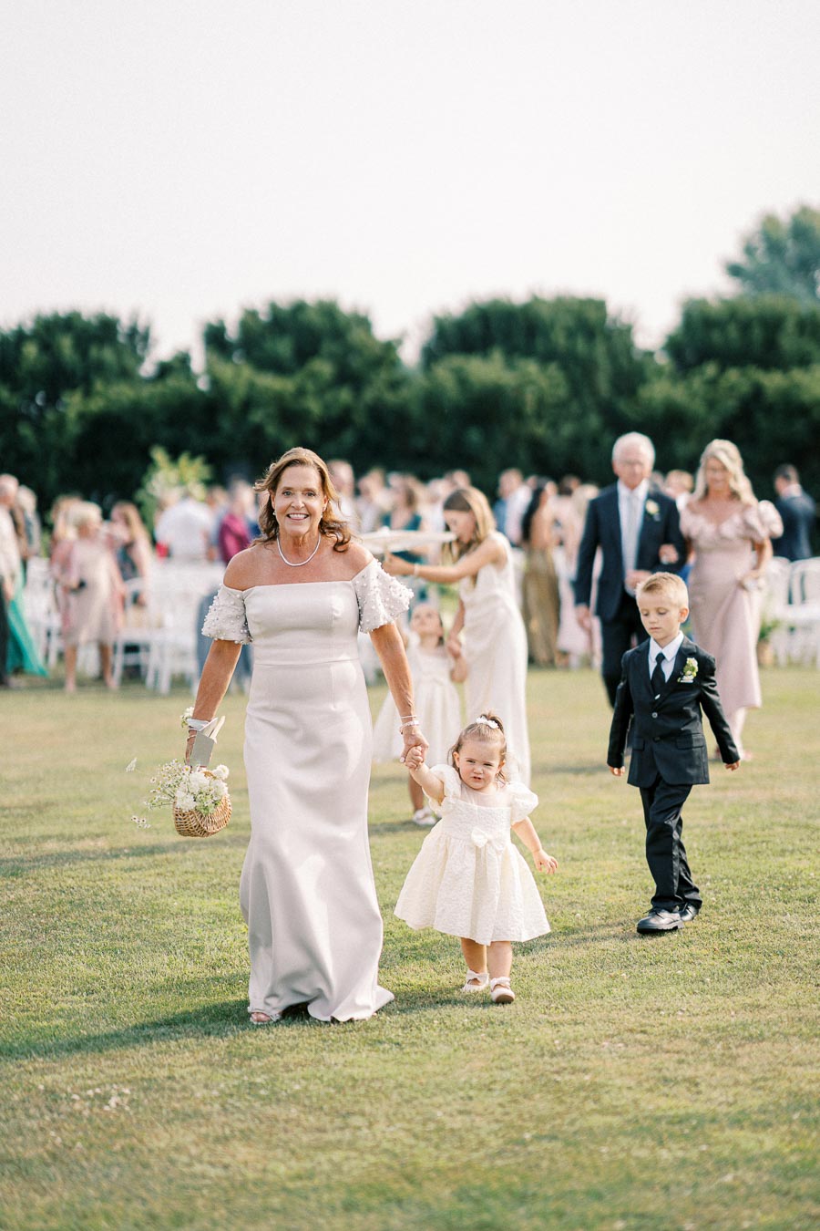 A woman in a white dress walks hand in hand with a young girl in a matching dress at an outdoor wedding ceremony, with guests and greenery in the background.