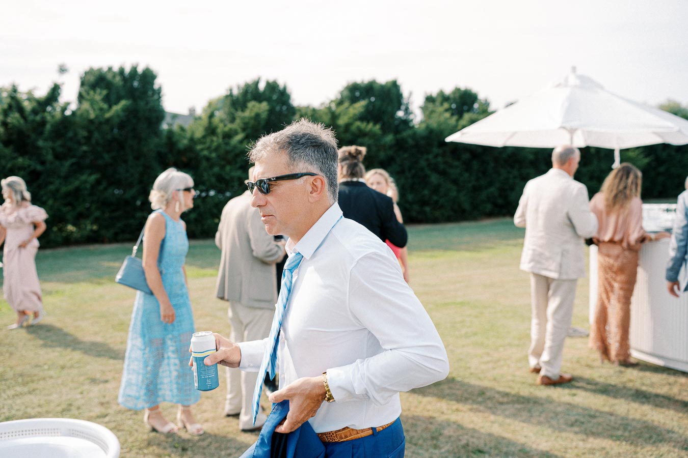 A man in sunglasses holds a beverage can while attending an outdoor gathering. He wears a white shirt and blue tie, with people socializing in the background near a large umbrella.