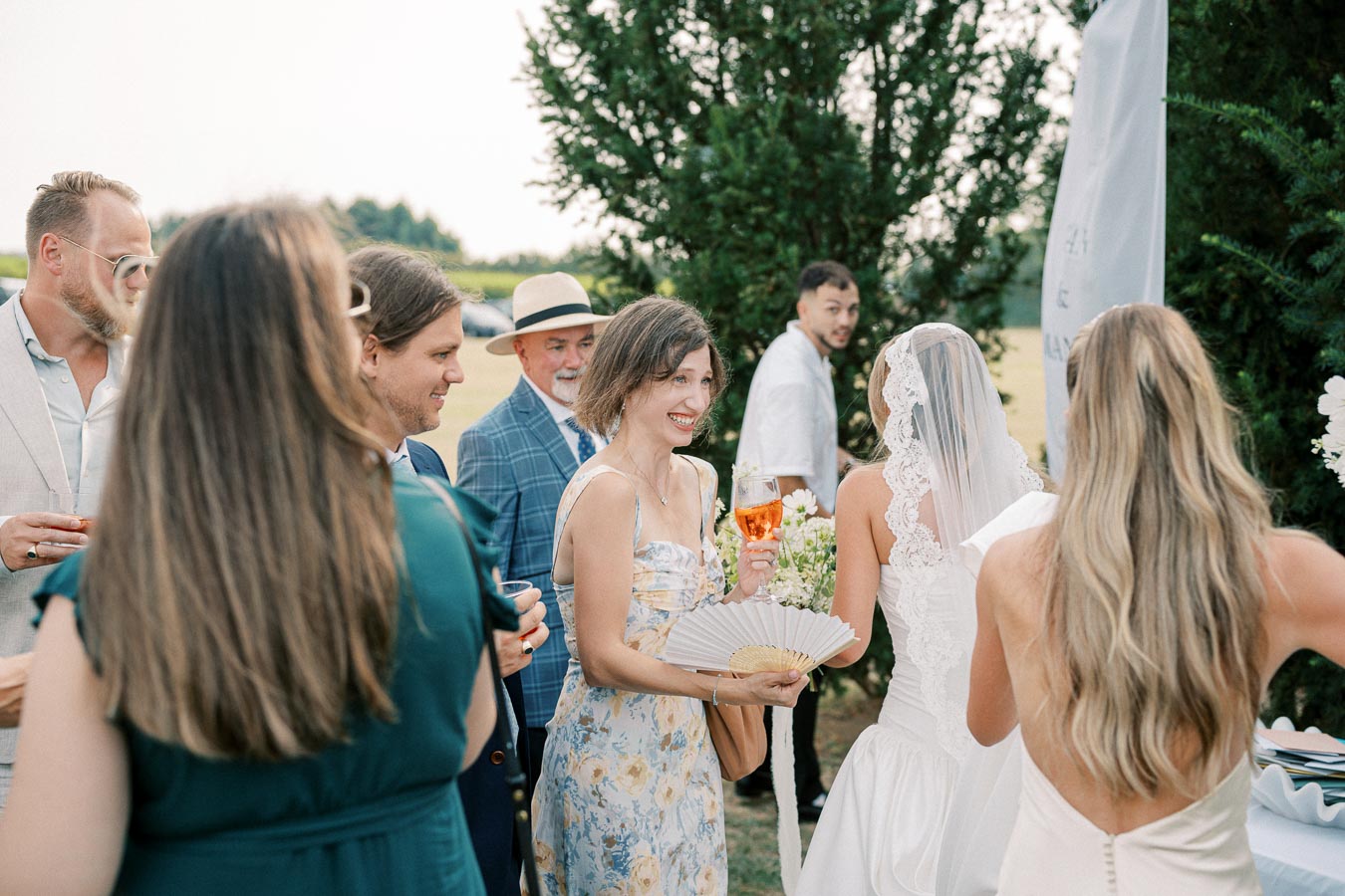 A group of well-dressed guests mingling outdoors at a wedding reception, with a bride in a lace veil, holding a bouquet and engaging with attendees in light summer attire, set against a backdrop of lush greenery.