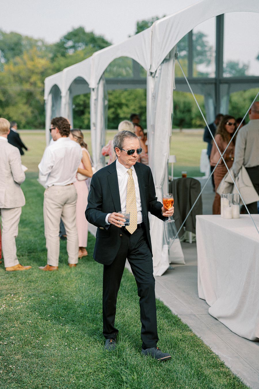 A man in a suit holding two drinks walks on a grassy lawn during an outdoor event with a white tent and guests in the background.