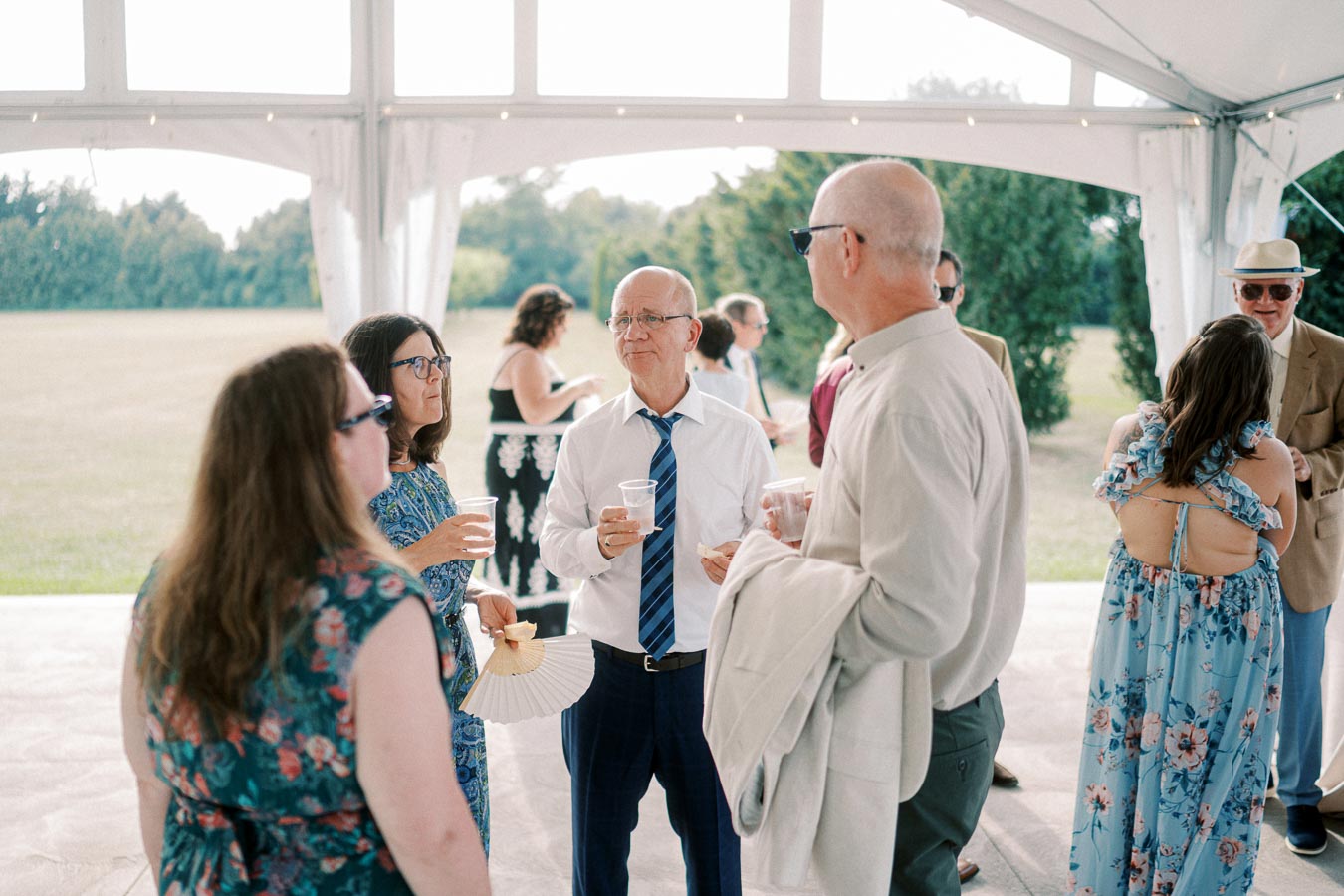 A group of people socializing at an outdoor event, standing under a white tent. They are dressed in summer attire, holding drinks, and conversing with each other. The background shows a field and trees, suggesting a relaxed, festive gathering.
