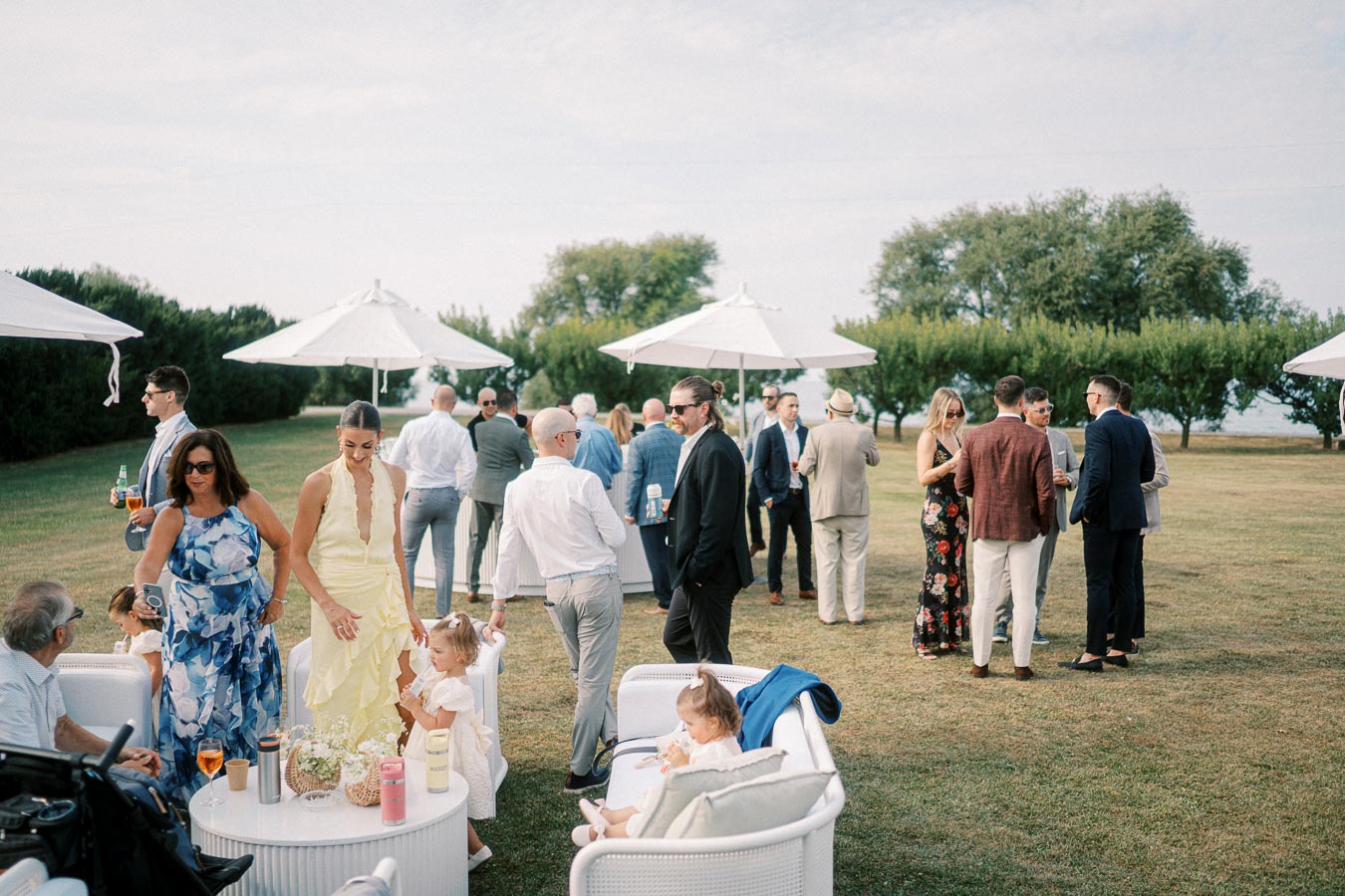 A group of elegantly dressed people socializing outdoors on a sunny day, with umbrellas providing shade and lush greenery in the background.